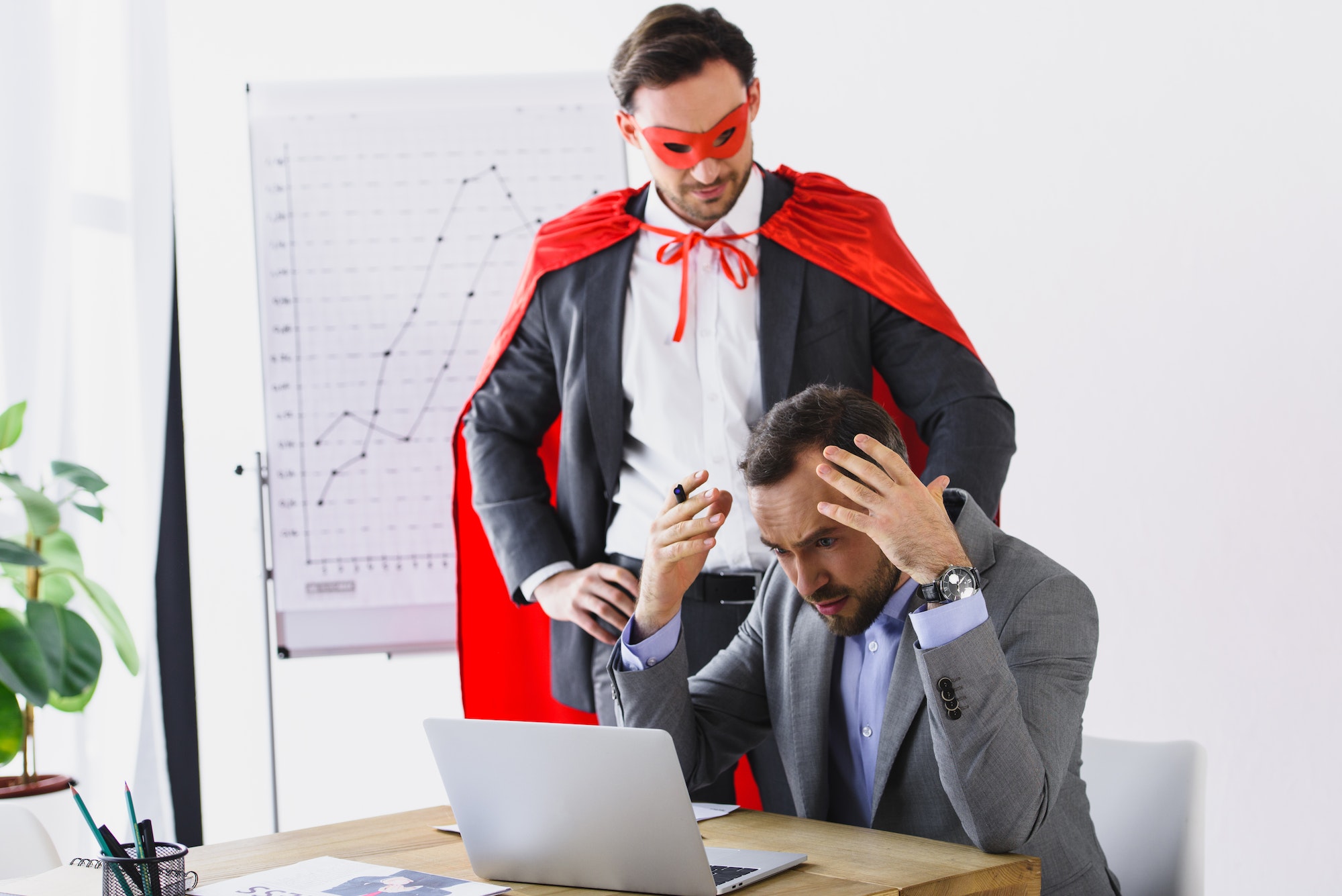 super businessman in mask and cape helping angry businessman with work in office